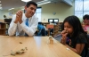 Matt Milosevich, a meteorologist with WLKY in Louisville, talks with 1st-grade student Jimena Jara as she experiments with a straw and building materials during a distance learning program at Lowe Elementary School. Milosevich talked about the effects of a 2008 wind storm in Louisville as part of the program, which was born out of a student's question about the strongest type of storm. Photo by Bobby Ellis, March 14, 2017