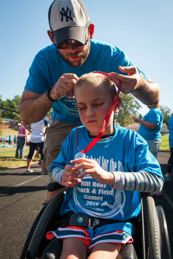 Kyle Sochia, a teacher at Kentucky School for the Blind, helps Colton Darnell, a third-grader at Kentucky School for the Blind, with his medal after his race. Photo by Bobby Ellis; Oct. 12, 2016