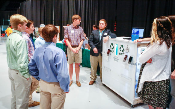 James Rogers, right center, a senior at the Early College and Career Center (Hardin County) explains a project created by students in the energy management program at the school to students from Anchorage Public Elementary School (Anchorage Indepenent) at the Kentucky Green and Healthy Schools and Kentucky National Energy Education Development (NEED) Project Youth Summit and Awards Luncheon at Frankfort Convention Center. Fellow Early College and Career Center seniors George Board, left center, and Alexa Johnson, right, listen as Rogers talks about a device that encourages people to recycle by providing 15 minutes of time to charge phones and other devices when they recycle a beverage container. Photo by Mike Marsee, April 27, 2017