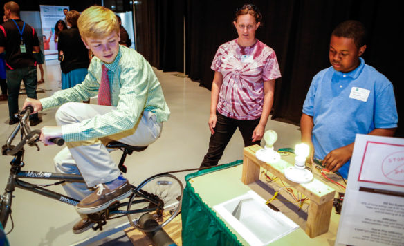 Ryan Burchett, an 8th-grade student at Anchorage Public Elementary School (Anchorage Independent) watches an LED bulb light up as a result of his activity on a stationary bicycle at an exhibit manned by Ockerman Middle School (Boone County) teacher Jennifer Davis and 8th-grade student Jordan Lowry. Photo by Mike Marsee, April 27, 2017