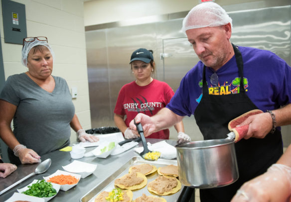 Mark MIller, a cook at Trimble County Schools, right, puts corn on Eagle Pizzas. Photo by Bobby Ellis, Aug. 1, 2018