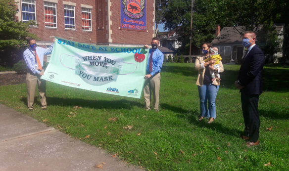 Three men and one woman stand in front of a school holding a flag with a face mask on it that says, "When you move, you mask."