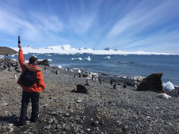 Donnie Piercey stands on a rocky beach with penguins and records a scene in Antarctica in 2018.