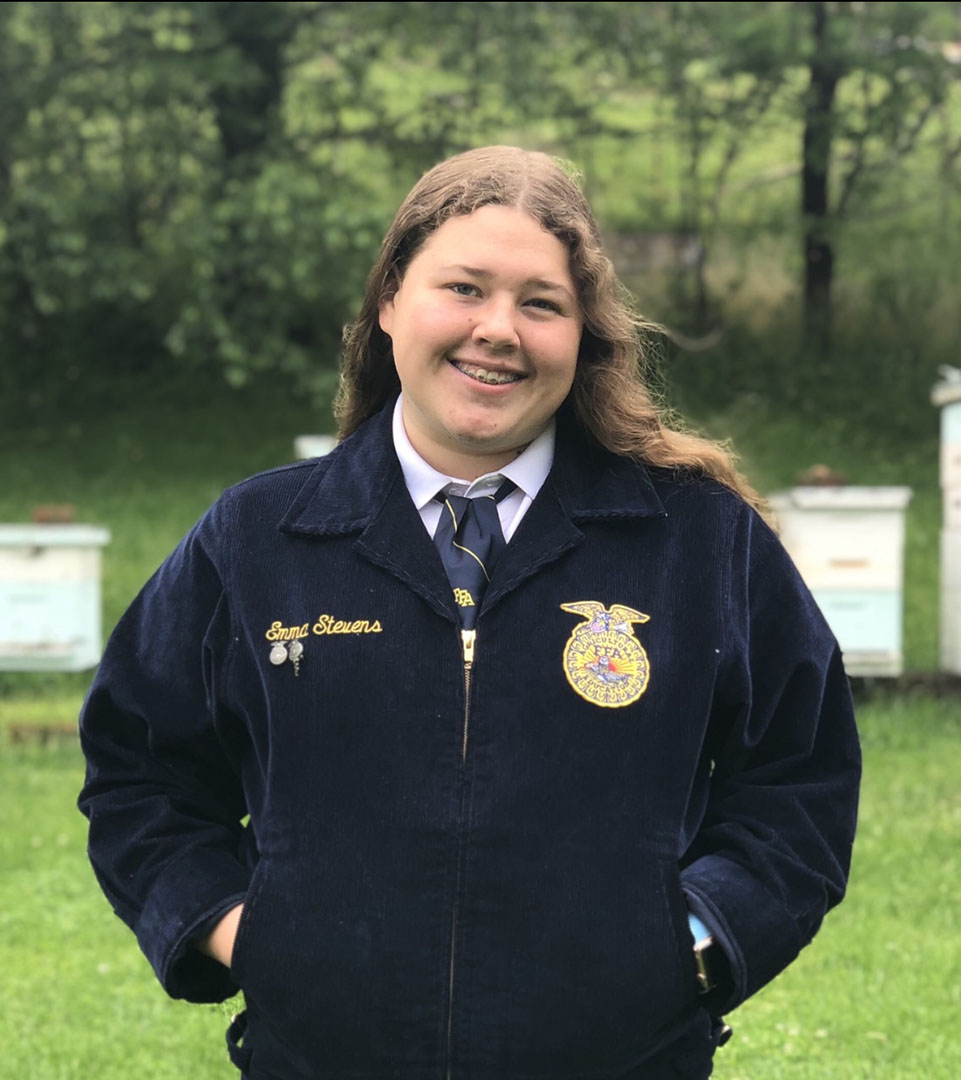 Picture of Emma Stevens wearing an FFA jacket and standing in front of bee hives.