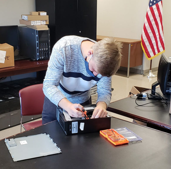 A photo of a young man wearing a face mask, bending over an opened computer laying on a table.