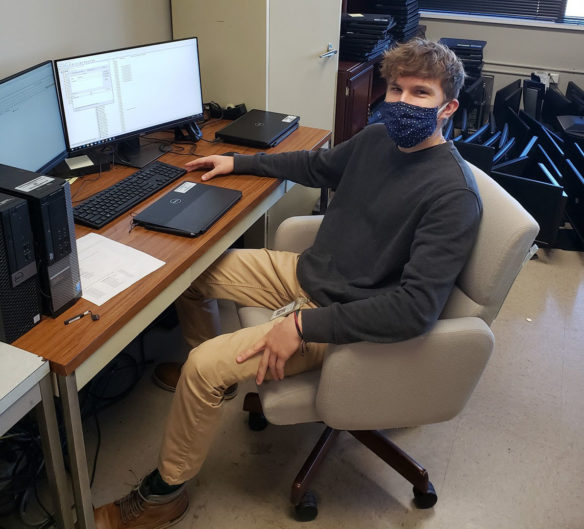 A picture of a young man wearing a face mask sitting down in front of a computer.