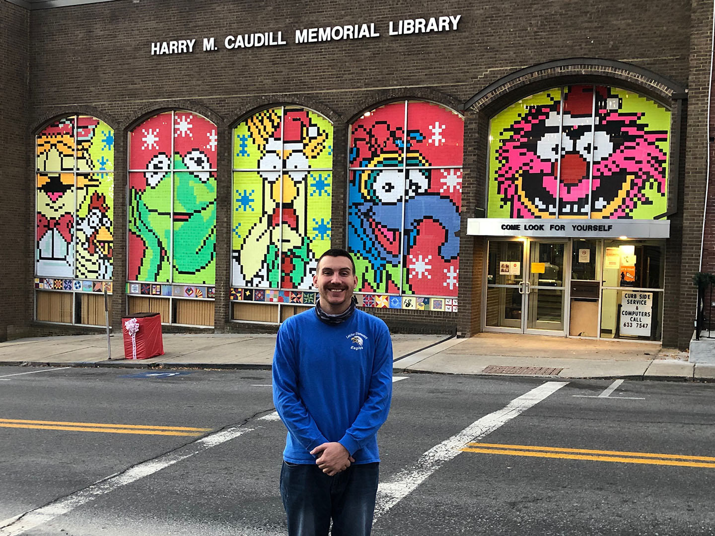 A man stands in front of the windows of a library that are decorated with brightly colored pictures of the Muppets.