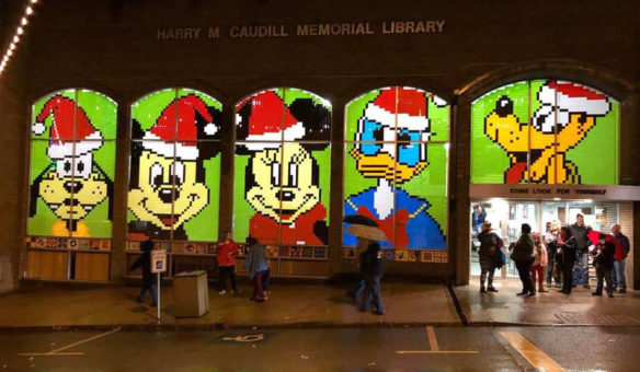 People stand outside of a library looking at windows decorated with brightly colored Micky Mouse characters.