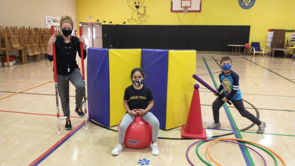 A picture of a woman on stilts, a girl sitting on a bouncing ball and a boy playing with a baseball bat in a gym.