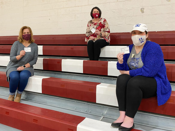 Three teachers sitting in bleachers, wearing face masks, hold up their vaccination cards.