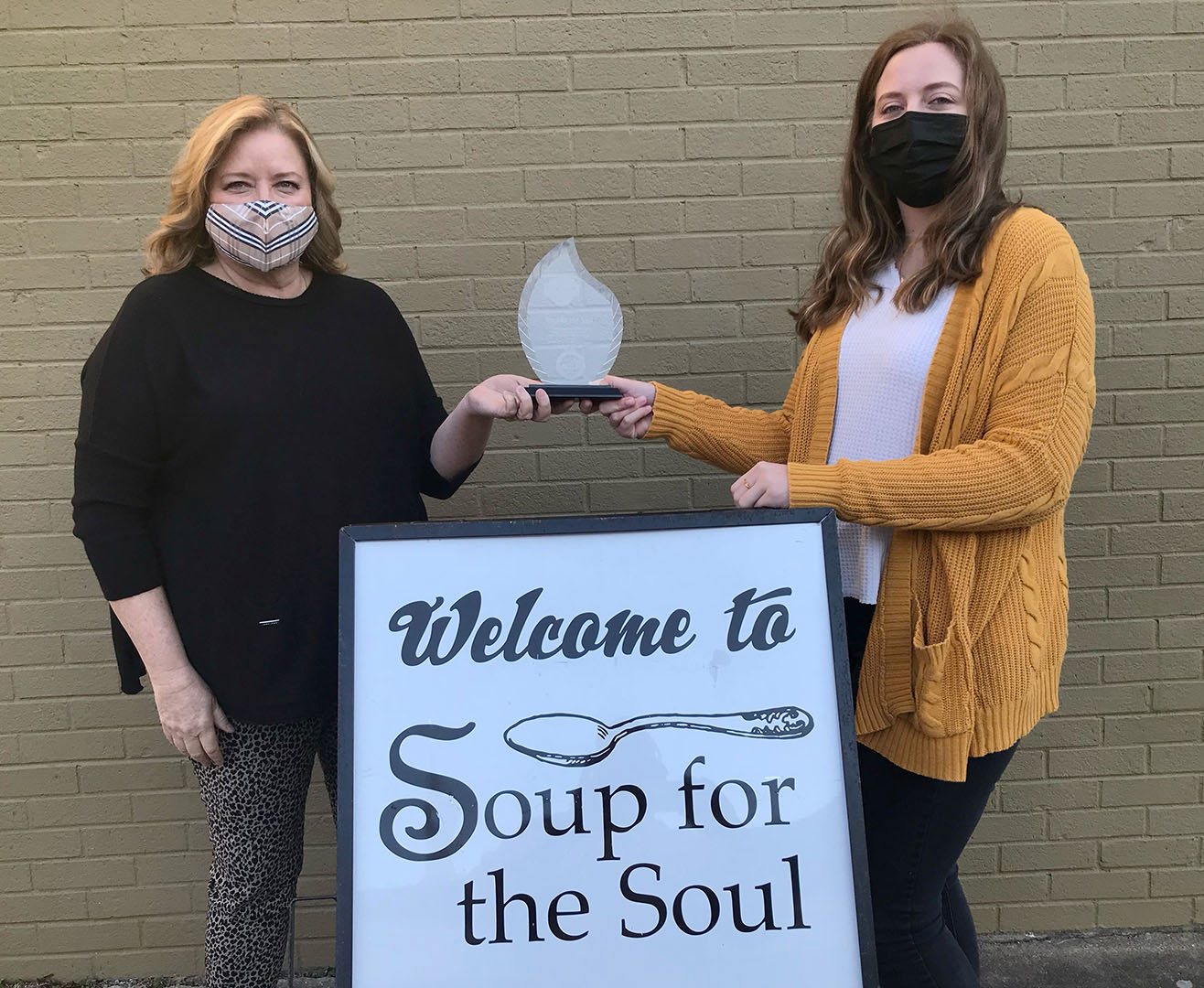 Two women wearing face masks hold onto a glass trophy over a wooden sign reading: Welcome to Soup for the Soul