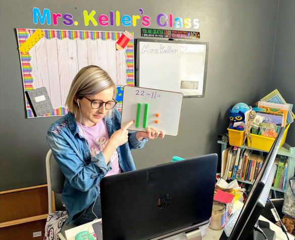 A woman sits in front of a computer, holding up a small sign with a math problem written on it.