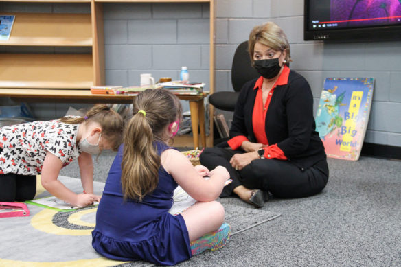 A woman sits on the floor talking to a young girl, while another girl colors nearby.