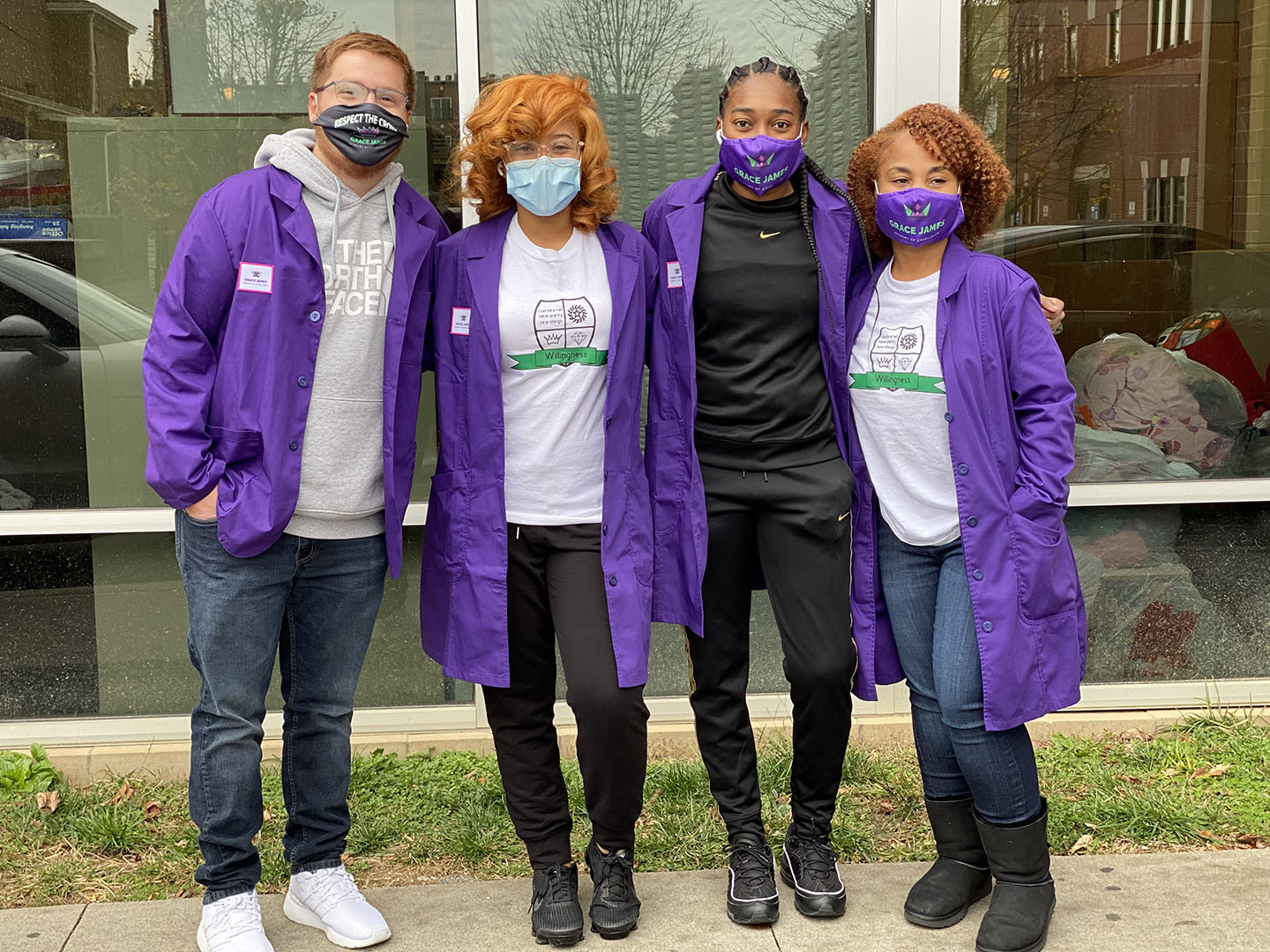 Picture of three women and one man, wearing facemasks and purple jackets, standing outside of a building with their arms around each other.