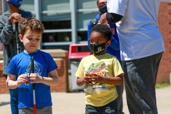 Two small boys stand next to each other. One holds a cane, the other is wearing a face mask.