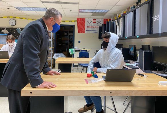 A man in a suit leans over a desk, talking to a male student sitting on a stool.