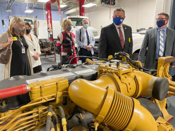 A group of people wearing faces masks tour a workshop with a diesel engine in the foreground.