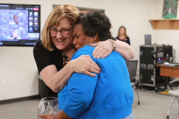 Picture of a smiling woman hugging another woman holding a glass vase.