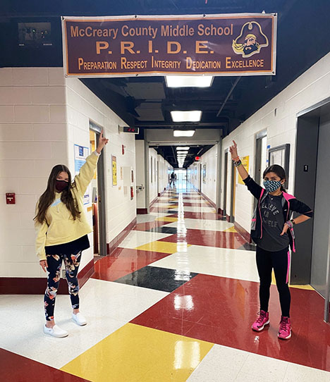 Two girls wearing face masks point to a banner hanging in the middle of a school hallway.