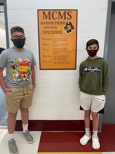 Two boys wearing face masks stand in a school hallway on either side of a sign titled MCMS Raider Pride Hallway Expectations.
