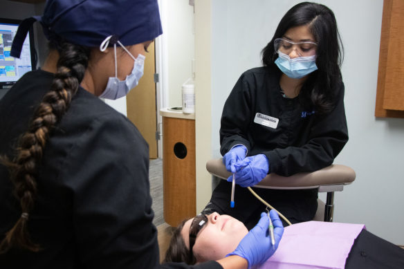 Two young women wearing facemasks stand over a patient laying back in a dental chair.