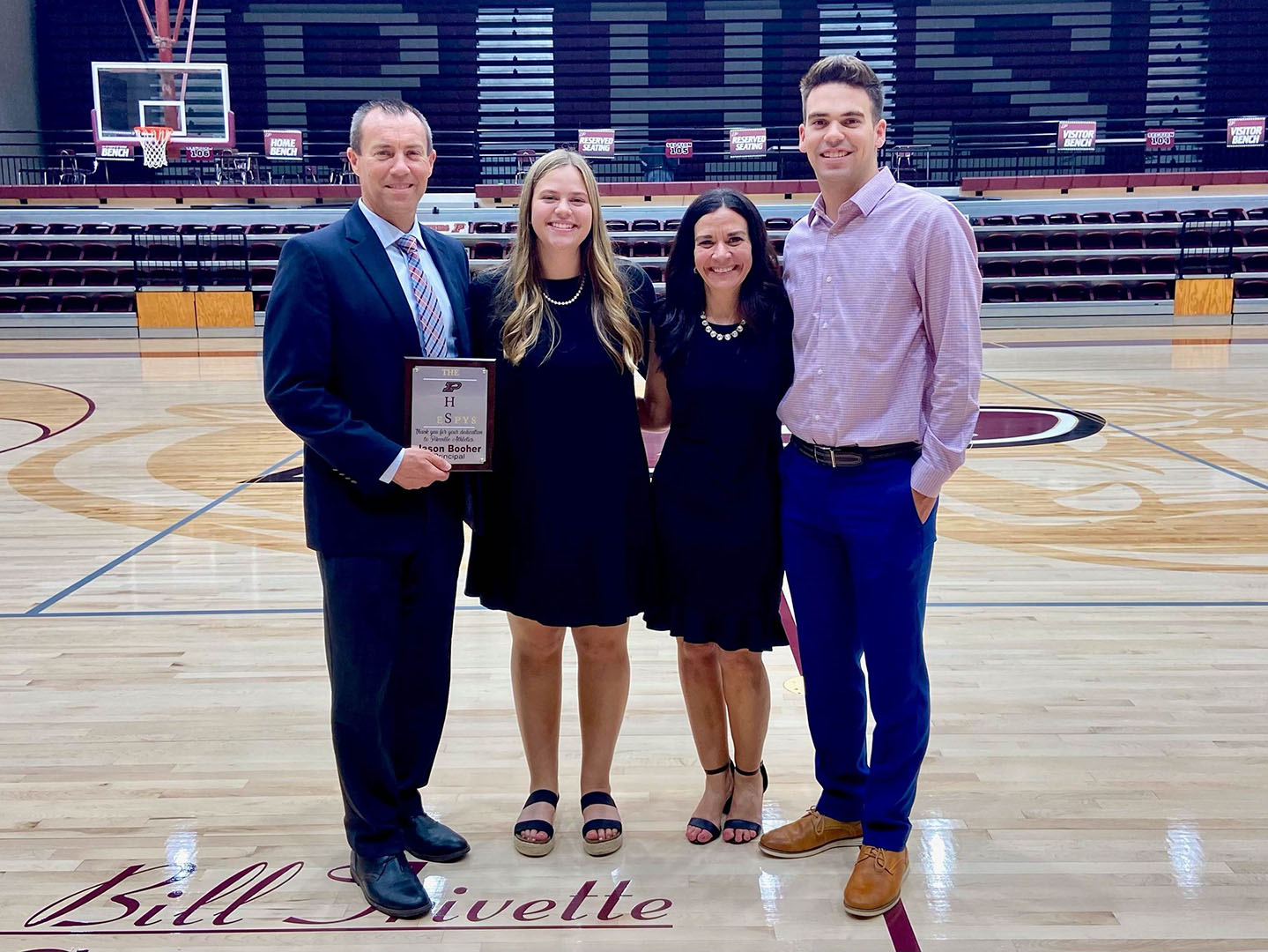 Picture of a man holding a plaque, two women and a young man standing on a basketball court, smiling.