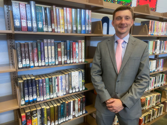 Picture of a smiling man in a suit standing in front of a row of books in a library.