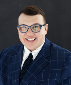 Picture of a smiling young man in a suit, wearing glasses.