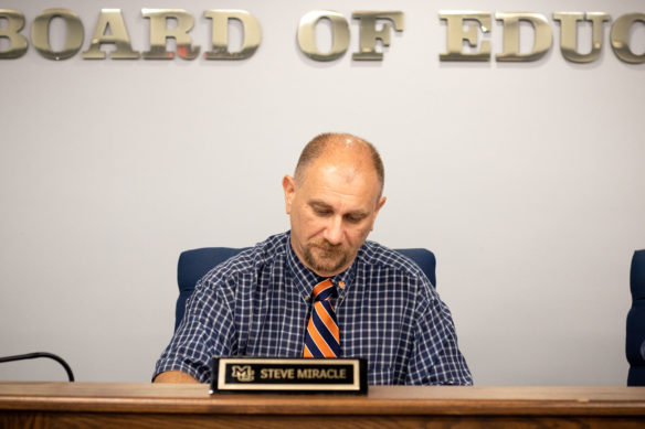 Picture of a man wearing a tie sitting behind a desk, with his eyes looking down at the tabletop.