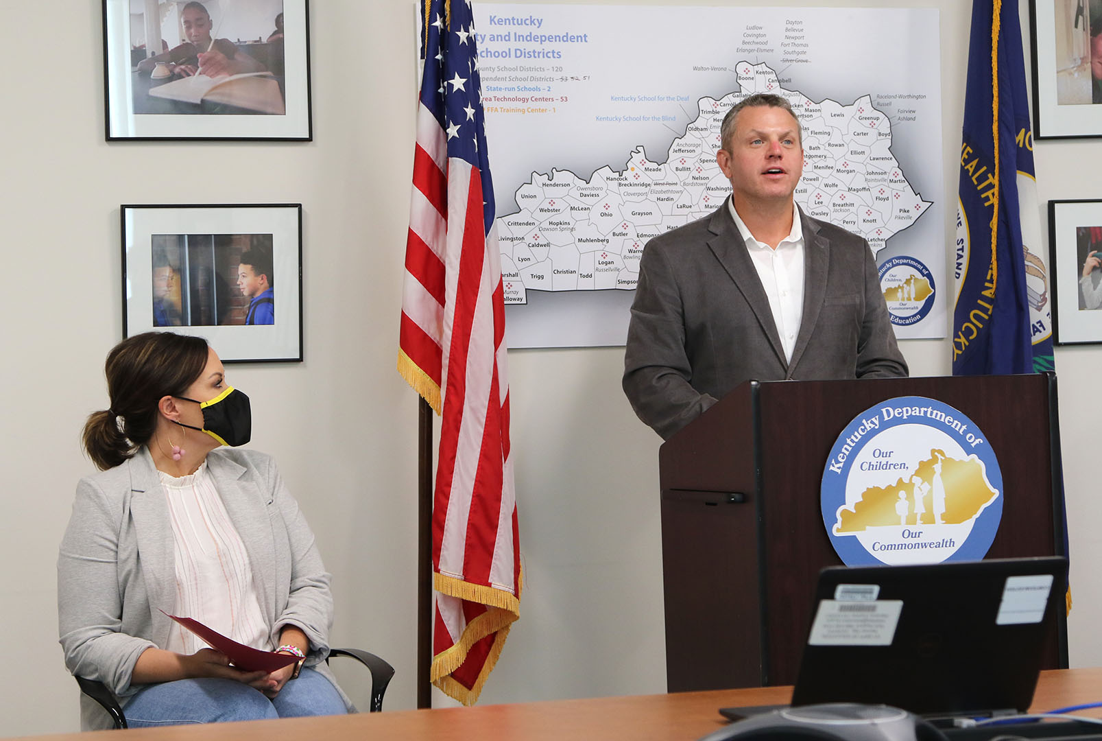 Picture of a man speaking at a podium, while a woman sits in a chair turned toward him.