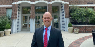 A picture of a smiling man, wearing a suit and standing in front of a school.