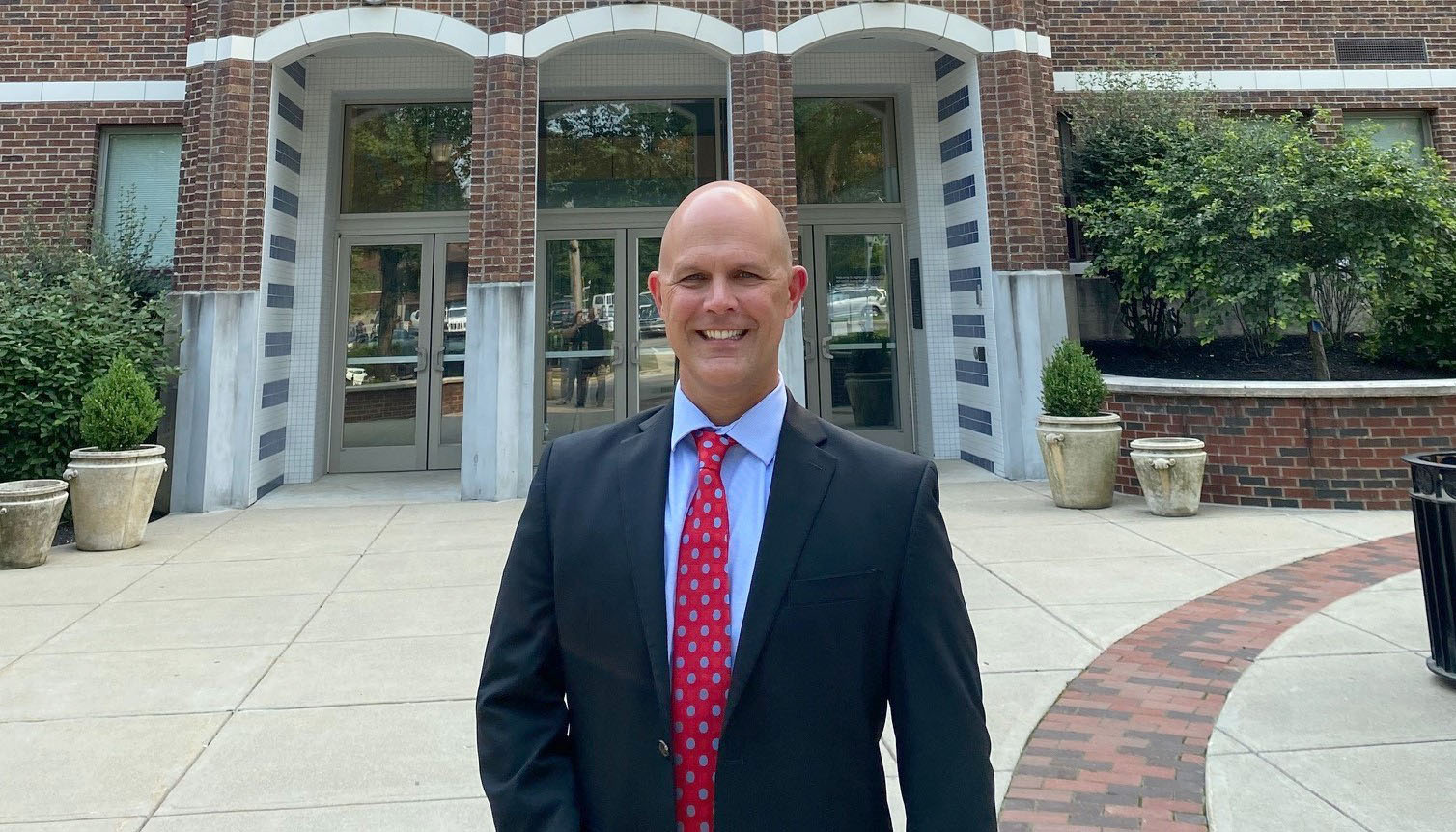 A picture of a smiling man, wearing a suit and standing in front of a school.