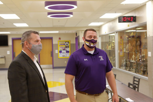 Two mean wearing face masks talk in a school hallway.