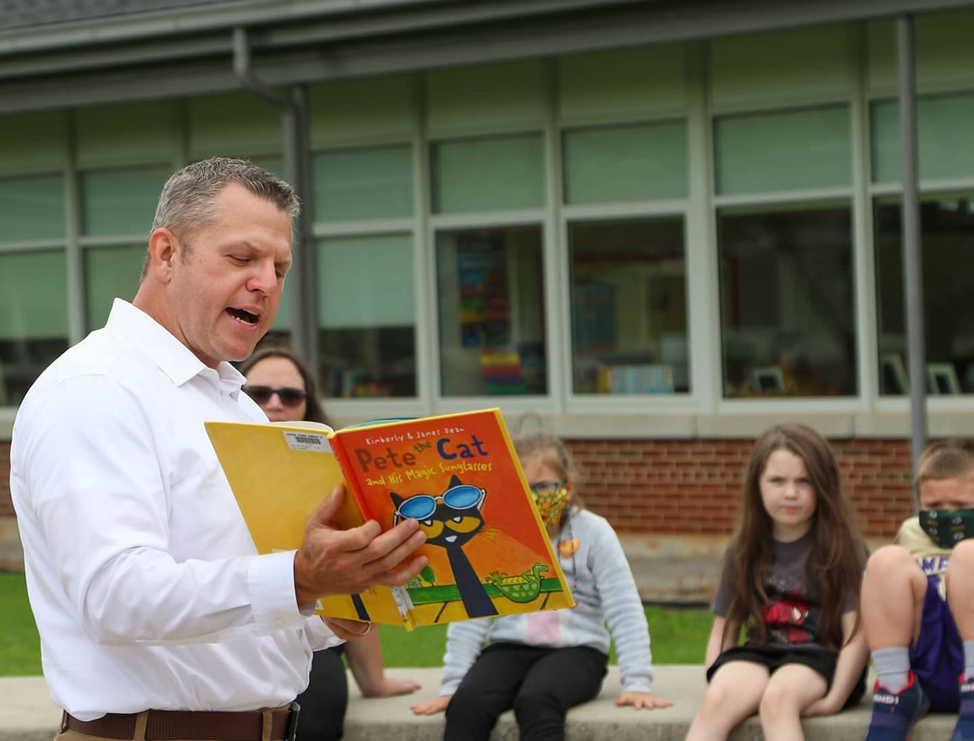 A man holding a book reads to children sitting outside.