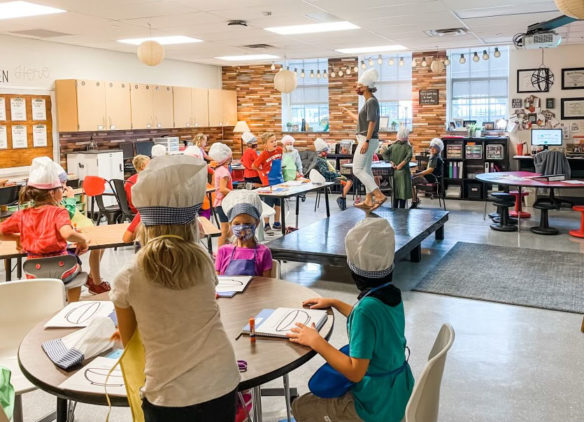 Picture of a woman wearing a chef's hat, standing on a small platform and talking to a room full of young children, who also wear chef's hats.