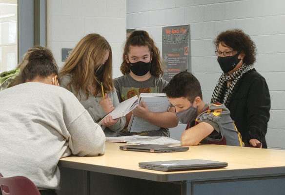 Picture of a woman standing behind a desk of students working together and looking over papers.