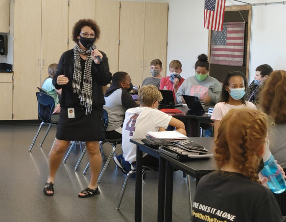 Picture of a woman talking into a microphone while students sit at their desks.