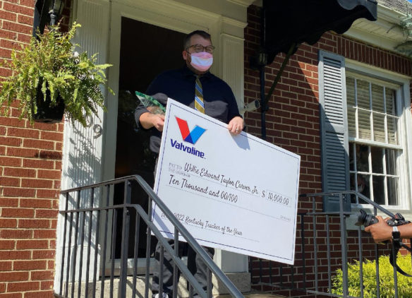 Picture of a man standing in front of his house holding a large presentation check and a glass award,.