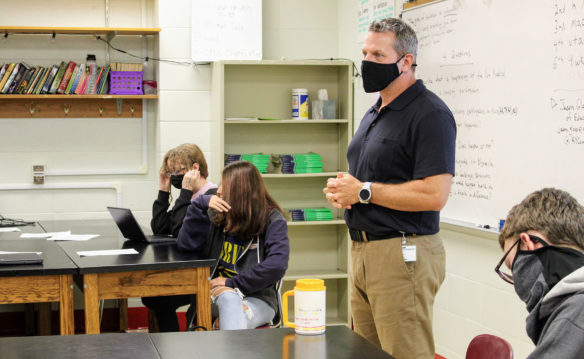 Picture of a man inside of a classroom wearing a face mask, talking in a classroom full of students.