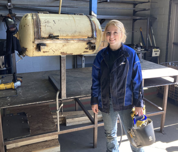 A young woman stands in front of a metal working table holding onto a welding visor and gloves.