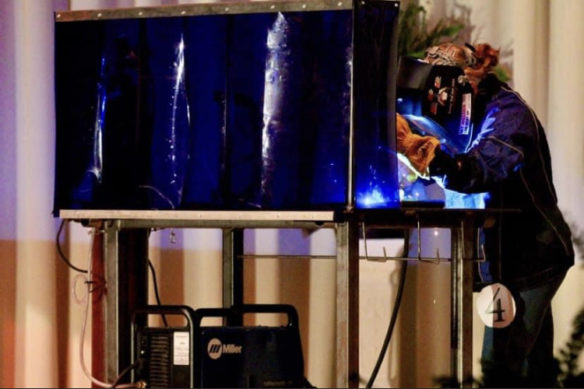 A woman stands on a stage behind a welding curtain, working with a torch. The number 4 hangs down from her jacket.