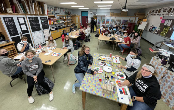 Wide picture of a room full of students sitting at tables with paint and brushes.
