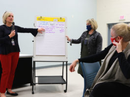 Picture of two women standing by a poster board with writing on it while talking to people in a large room.