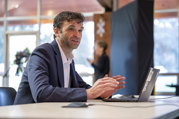 Picture of a man wearing a suit, sitting at a table and talking.