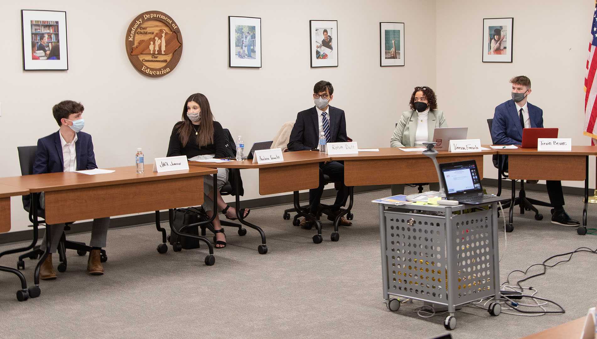 A picture of five high school students, wearing face masks, sitting behind a long conference room table and having a discussion.