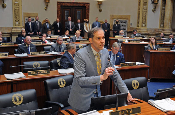 Picture of a male legislator standing at his desk, speaking, while people stand in the back of the room.