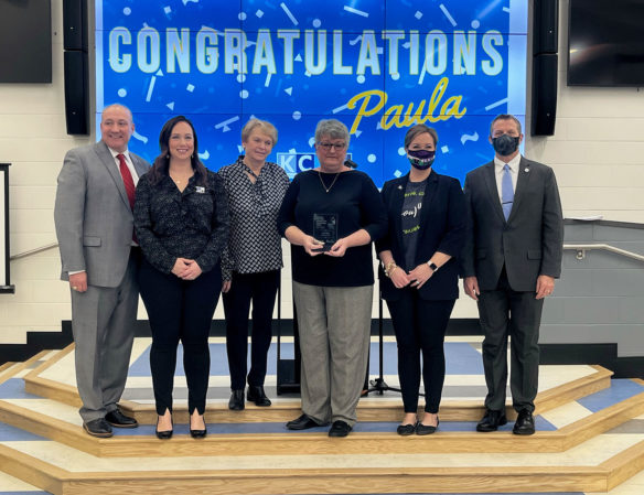 Picture of six people standing in front of a screen that reads Congratulations Paula. The woman at the center is holding a glass trophy.