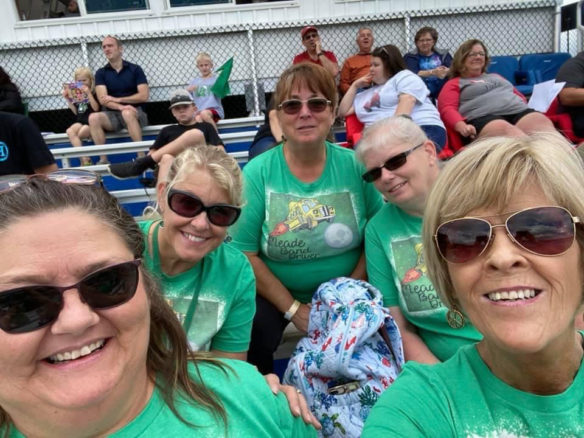 Five women sitting in the bleachers at an athletic game wearing sunglasses and smiling. 