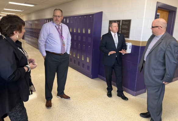 Three men and one woman stand in a high school hallway talking with lockers behind them.