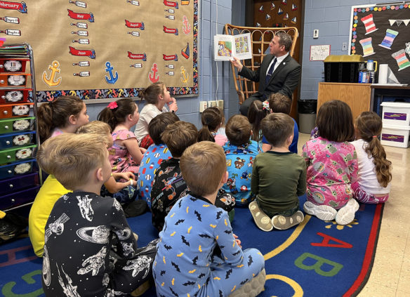 A man reads a children's book to a group of students sitting on a rug in a classroom.